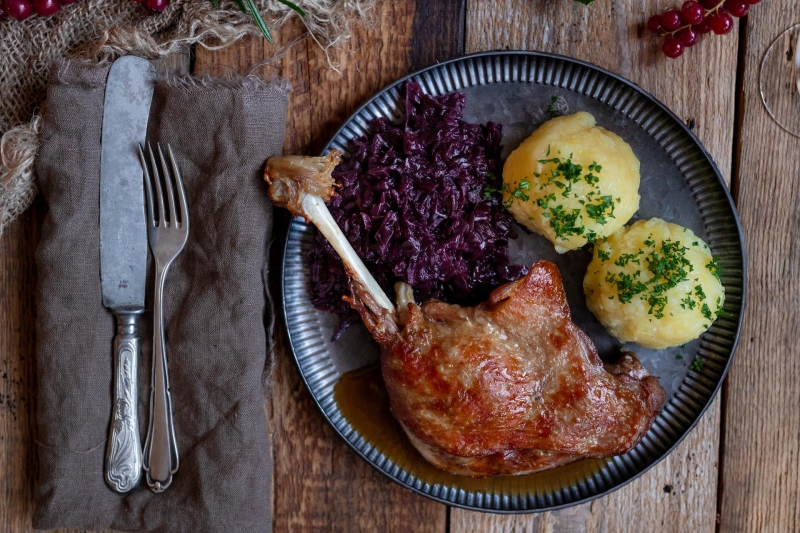 Gänsekeule mit Rotkohl, Klößen und dunkler Sauce. Auf dunklem Teller, daneben Besteck. Untergrund aus Holz.