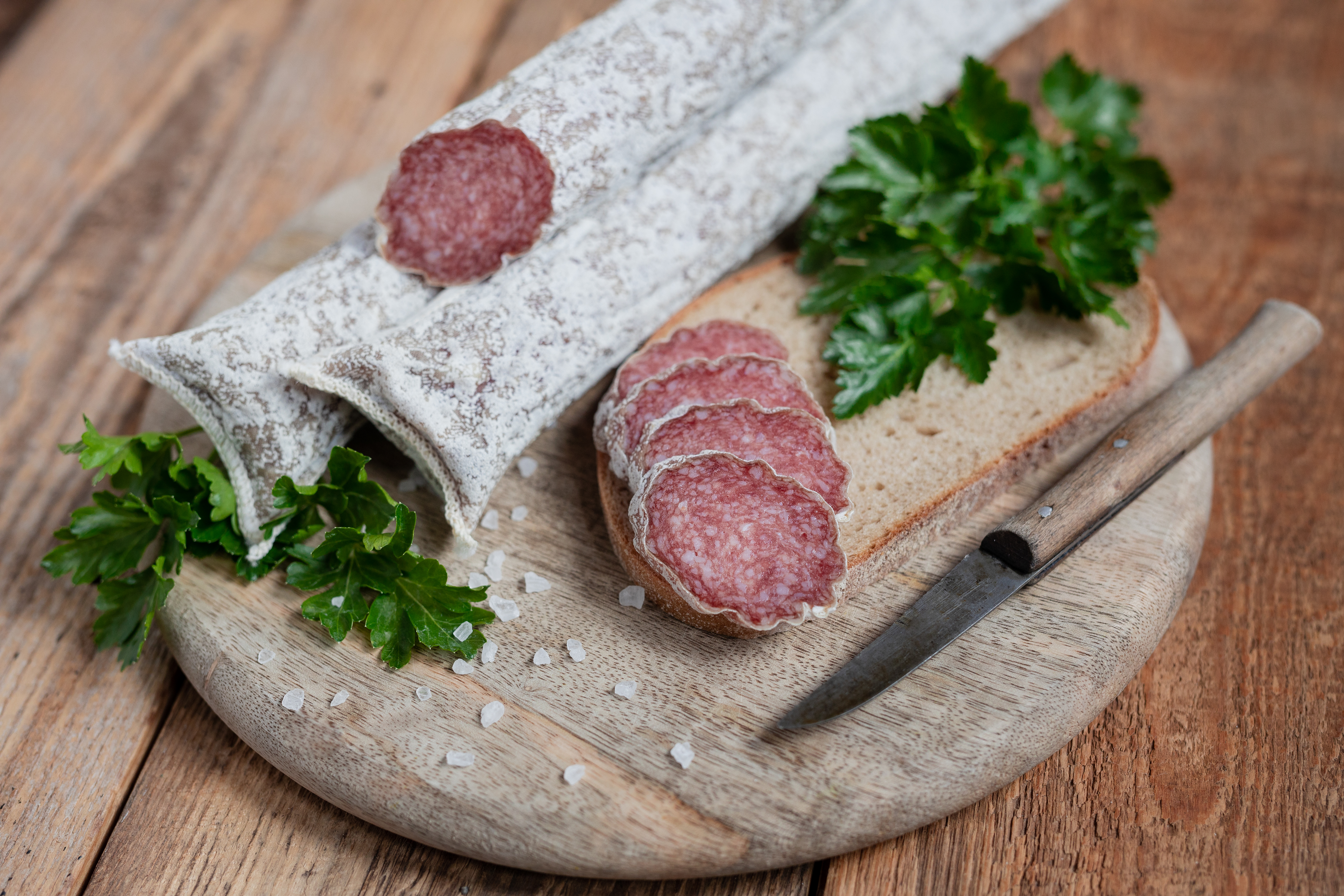 Auf einem Brett zwei mediterrane Salami. Daneben drei Scheiben der Salami, Kräuter und ein Messer.