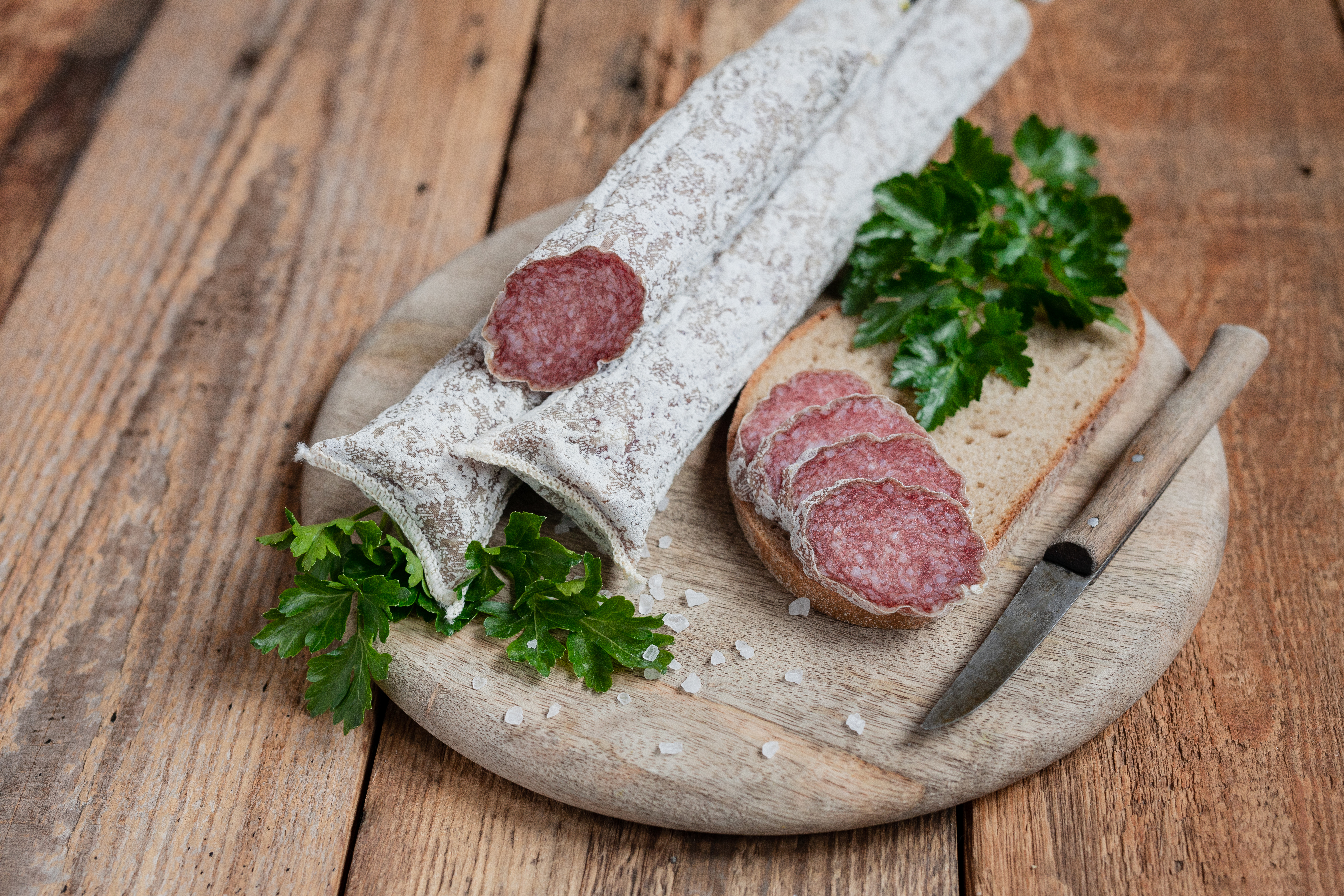 Auf einem Brett zwei mediterrane Salami. Daneben drei Scheiben der Salami, Kräuter und ein Messer.