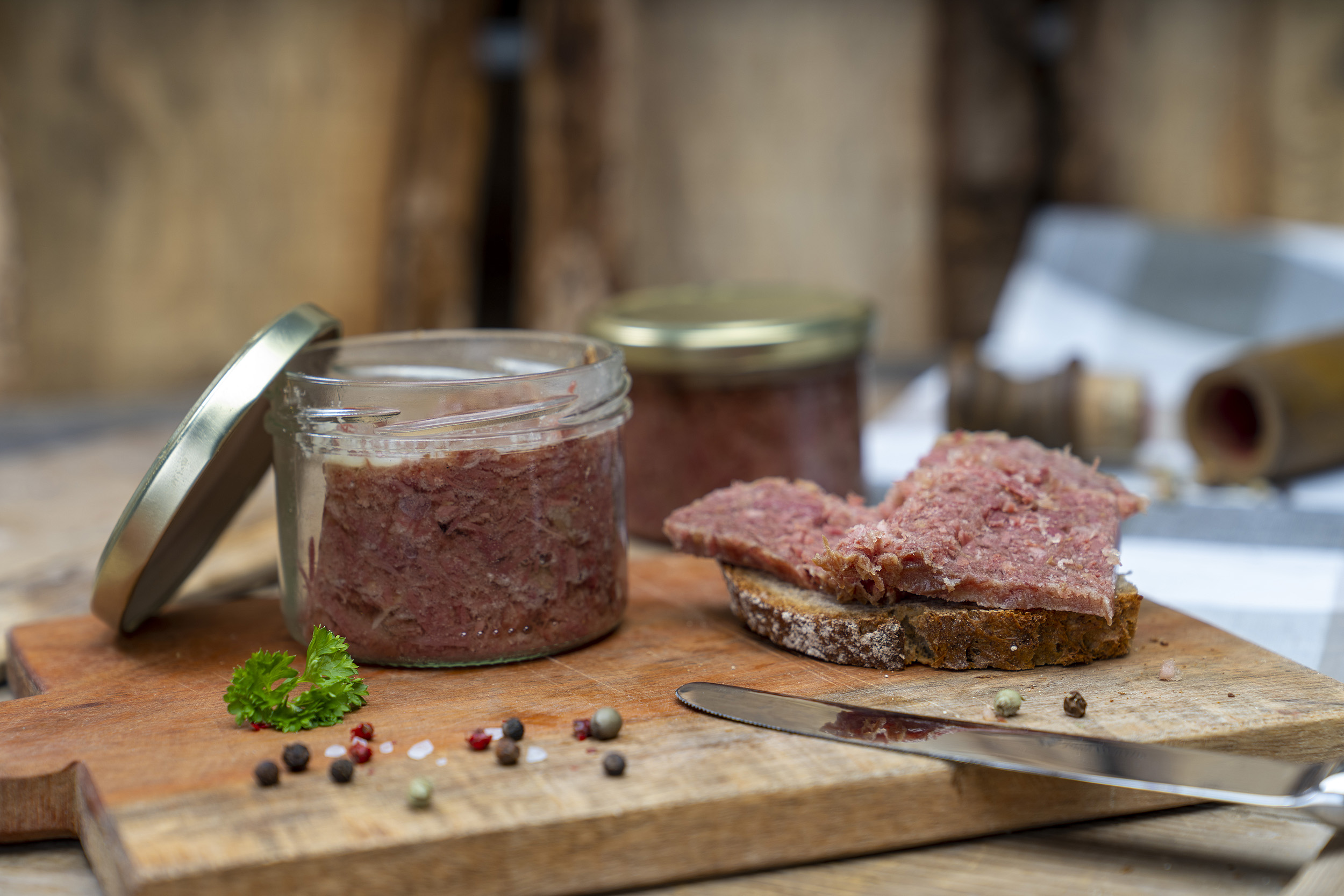 Auf einem Brett ein Glas Corned beef. Daneben ein geschmiertes Brot mit Corned beef. Daneben Gewürze, Kräuter und ein Messer. Im Hintergrund ein weiteres Glas.
