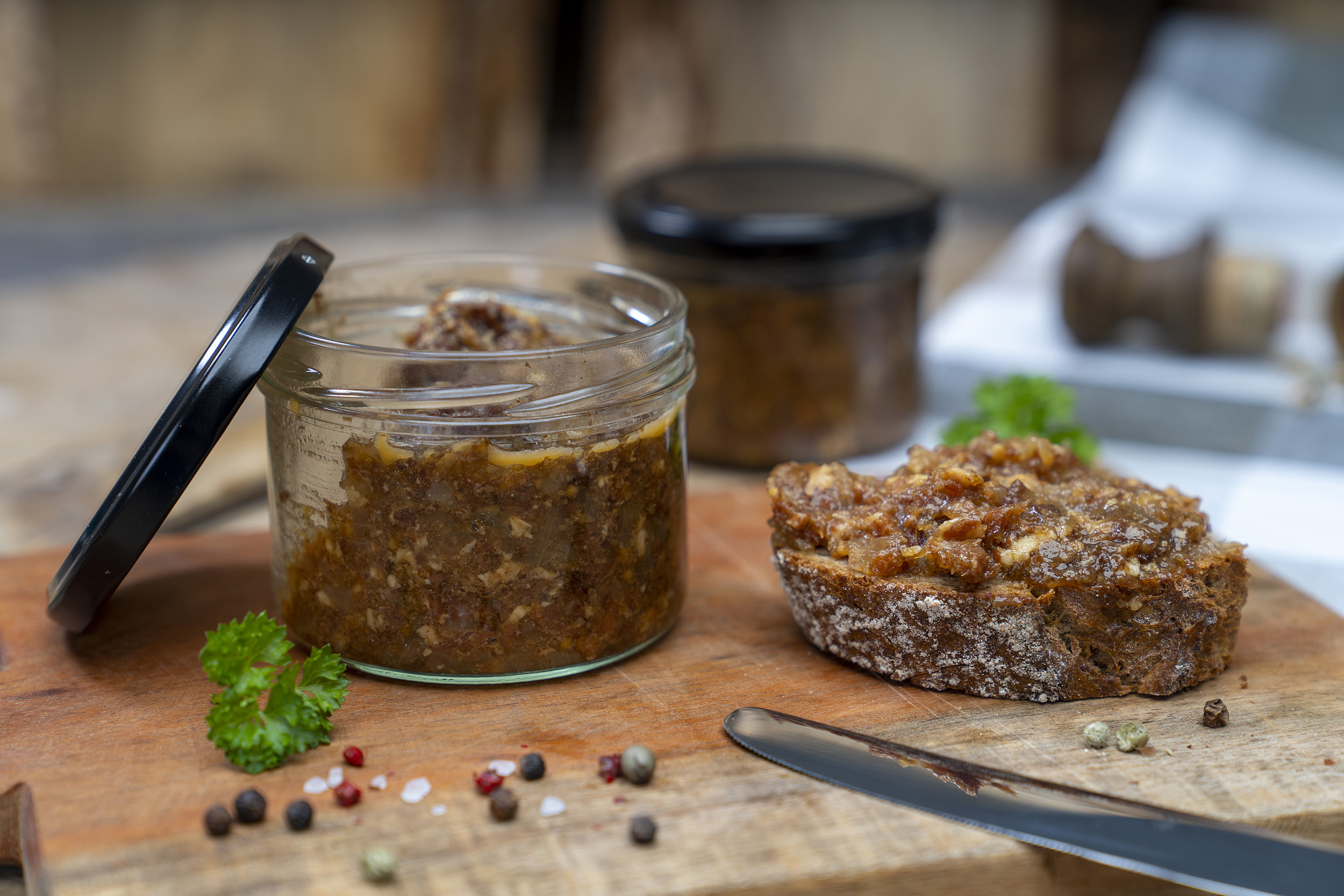 Auf einem Holzbrett steht ein Glas mit Bacon Jam. Daneben Gewürze, Kräuter und ein geschmiertes Brot mit der Marmelade. Im Hintergrund ein weiteres Glas. Vorn ein Messer am Brett.