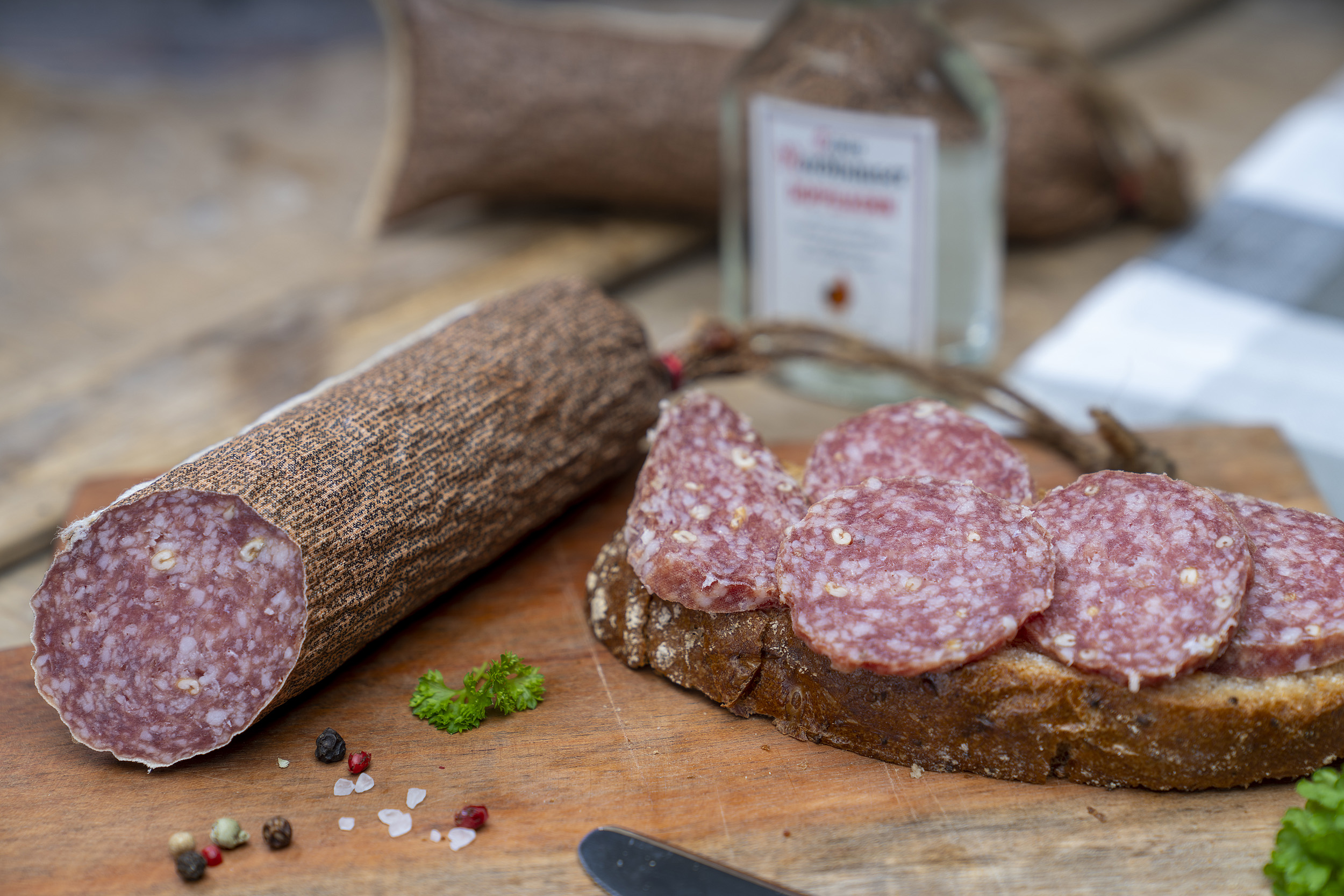 Kornsalami auf einem Holzbrett. Daneben eine Scheibe Brot mit der Salami und Gewürze. Im Hintergrund eine kleine Flasche Echter Nordhäuser Doppelkorn.