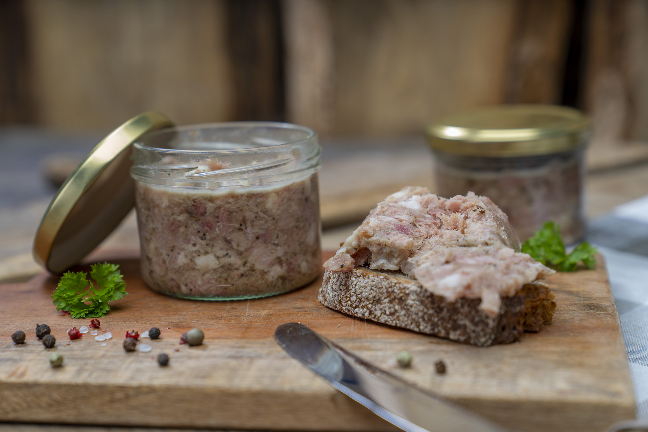 Auf einem Brett ein Glas Sülze. Daneben eine Schnitte mit der Wurst. Drum herum Kräuter und hinten noch ein Glas der Wurst.