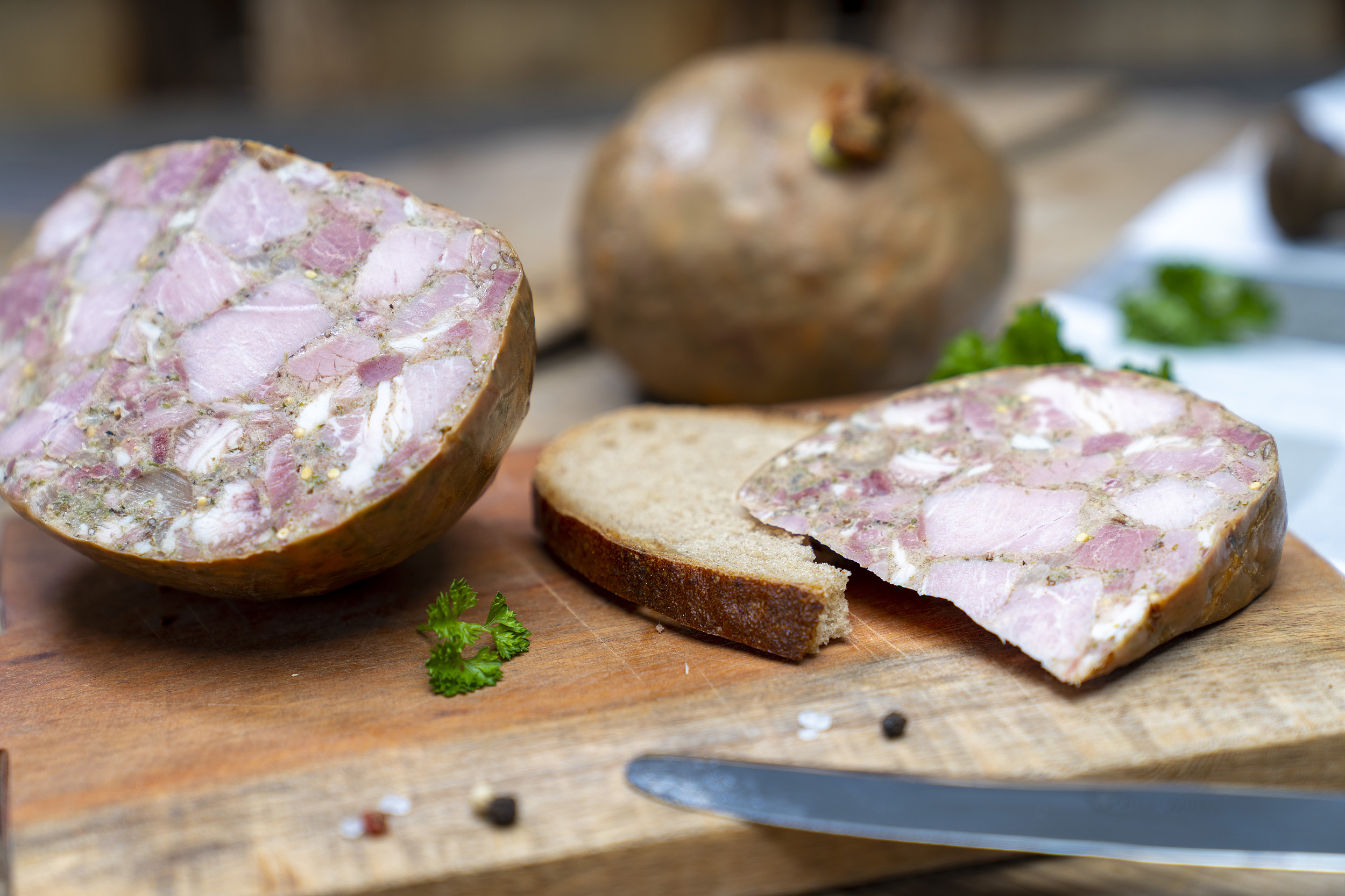 Eine Scheibe mit Sülze auf einem Brett. Daneben die angeschnittene Blase der Wurst und Gewürze. Hinten eine weitere Blase.