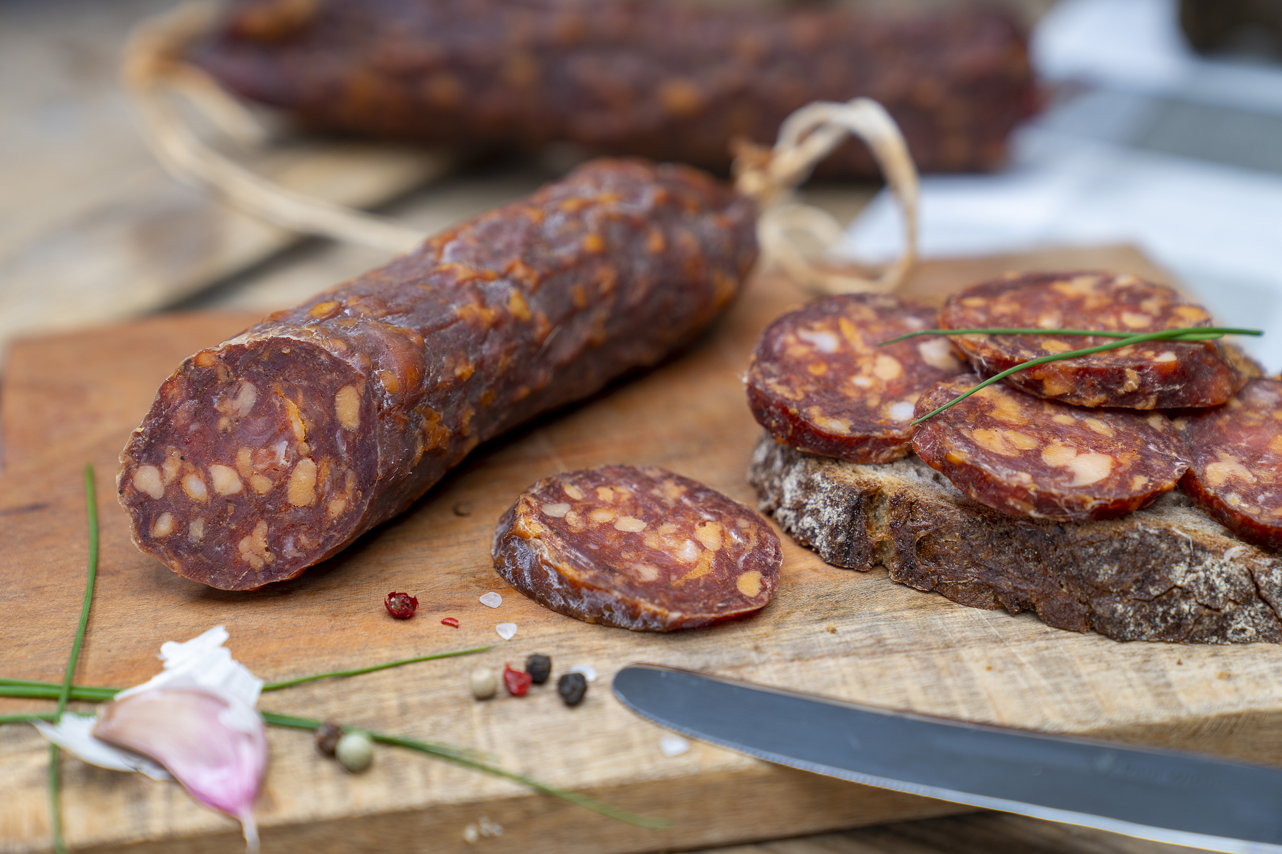 Angeschnittene Ungarische Salami auf einem Brett. Daneben eine Schnitte mit der Wurst und Gewürze. Im Hintergrund eine weitere Salami.