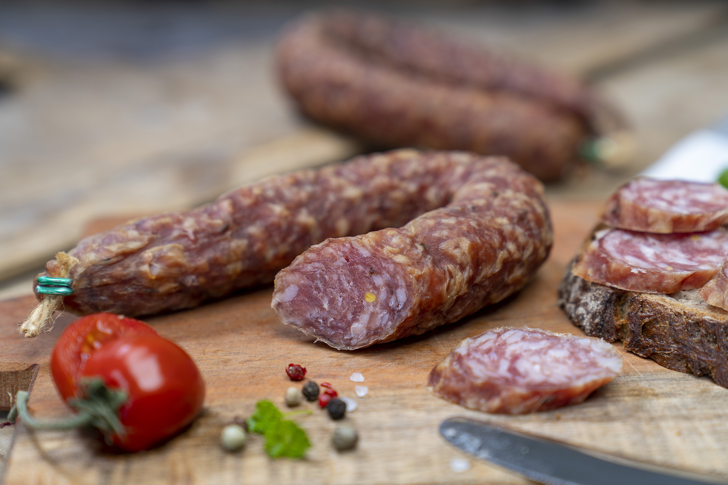 Ein Südharzer Knüppelchen auf einem Brett. Daneben ein Brot mit der Wurst, Gewürze und eine Tomate.