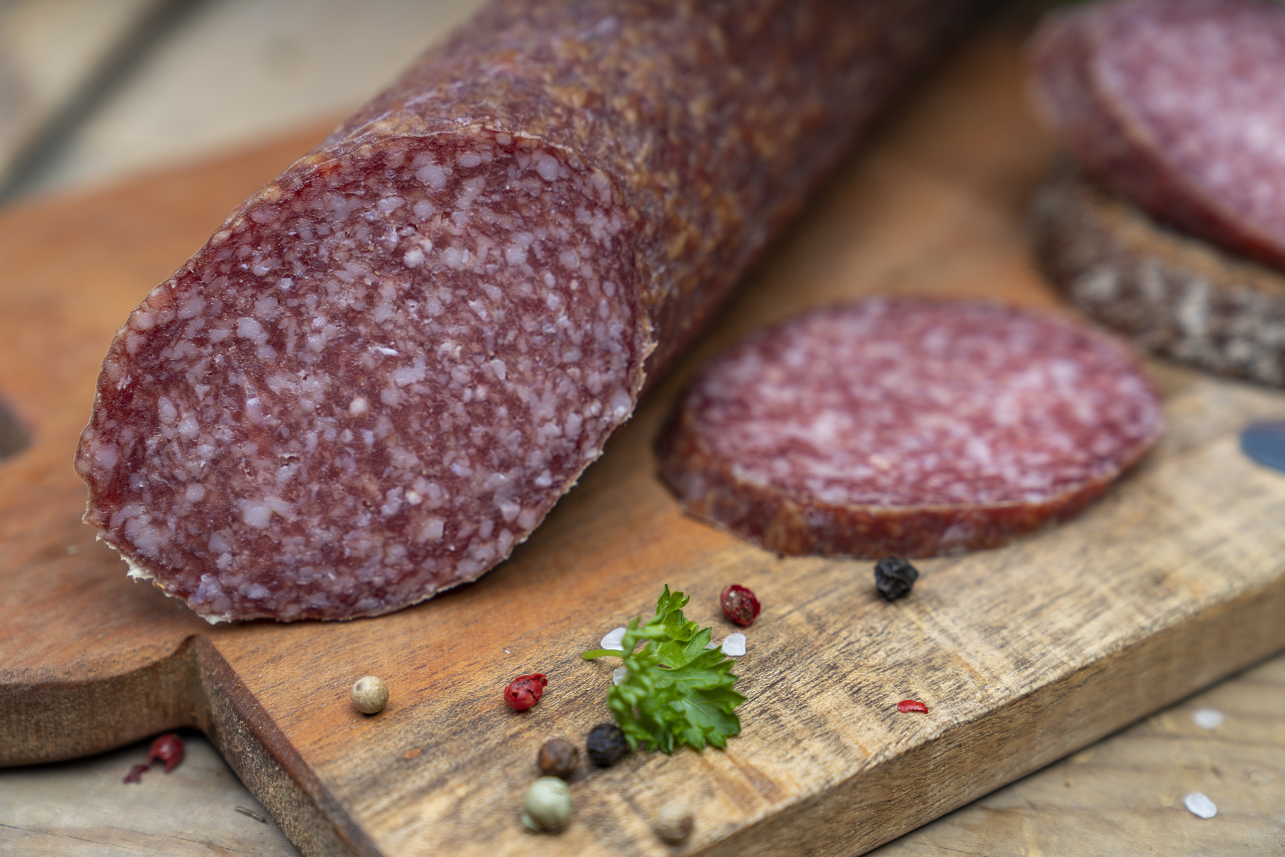 Nahaufnahme einer Bauernsalami. Daneben Kräuter und eine Scheibe.