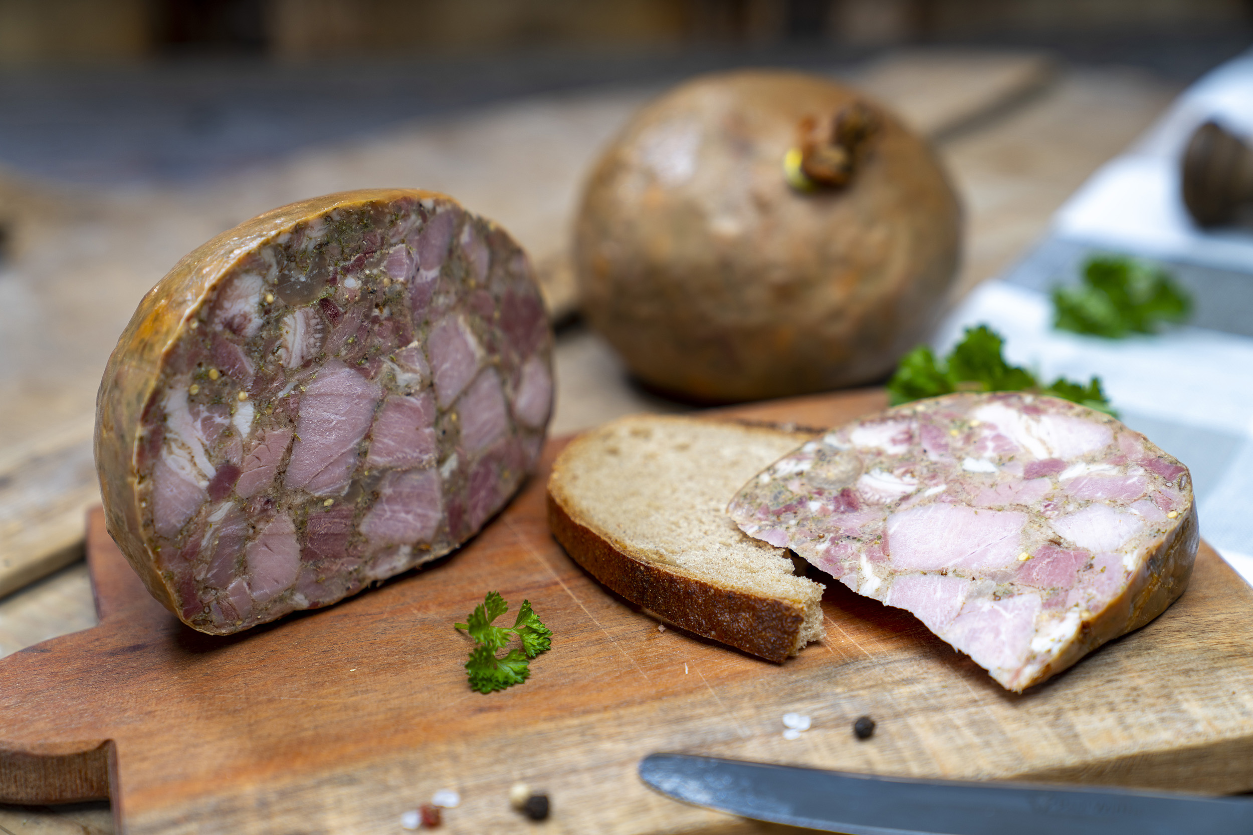 Eine Scheibe mit Sülze auf einem Brett. Daneben die angeschnittene Blase der Wurst und Gewürze. Hinten eine weitere Blase.