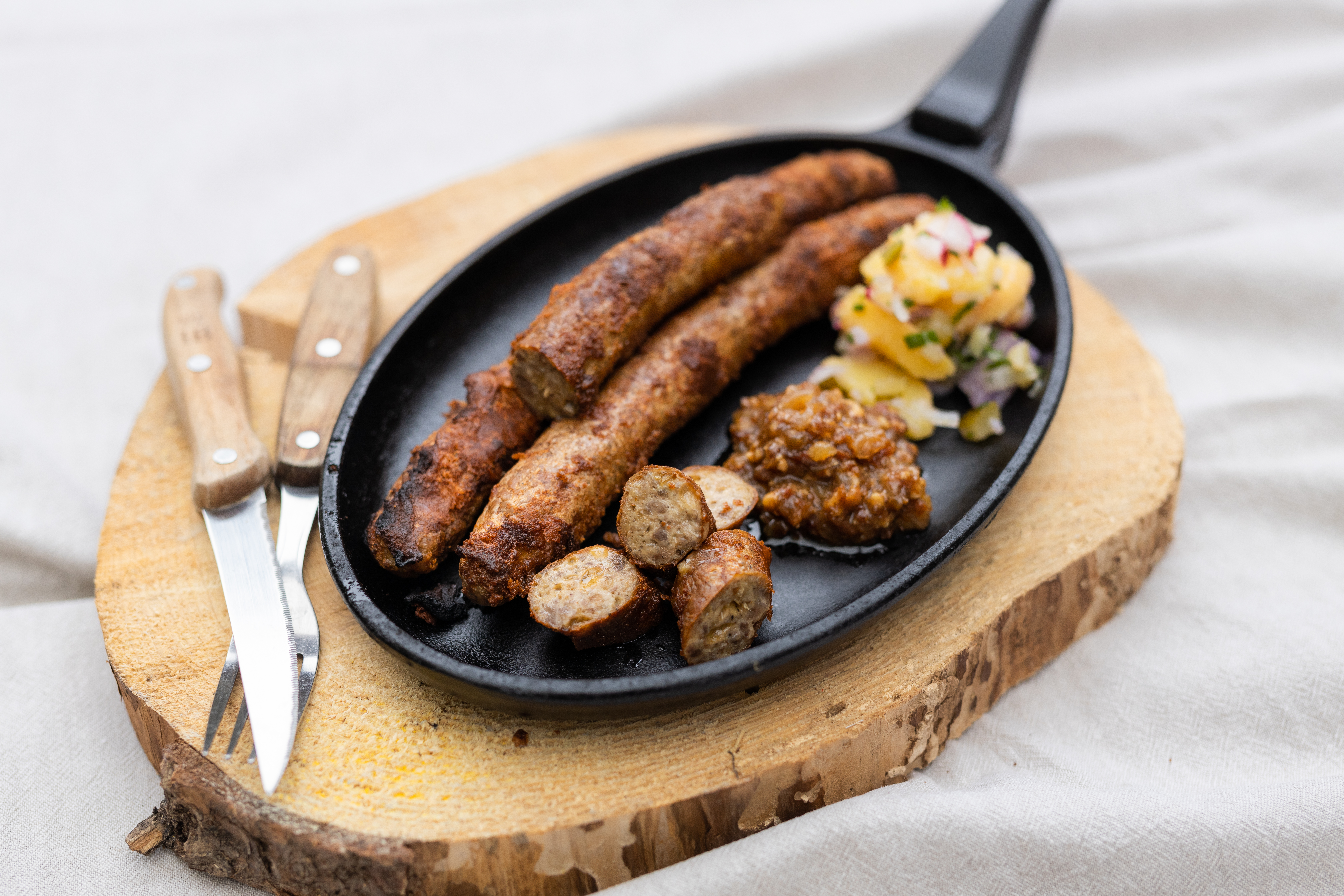 Drei Cheesburger Bratwürste in einer Pfanne. Einige Stücke sind abgeschnitten. Daneben Saucen und Salatbeilage. Unter der Pfanne ein Brett und Besteck daneben.