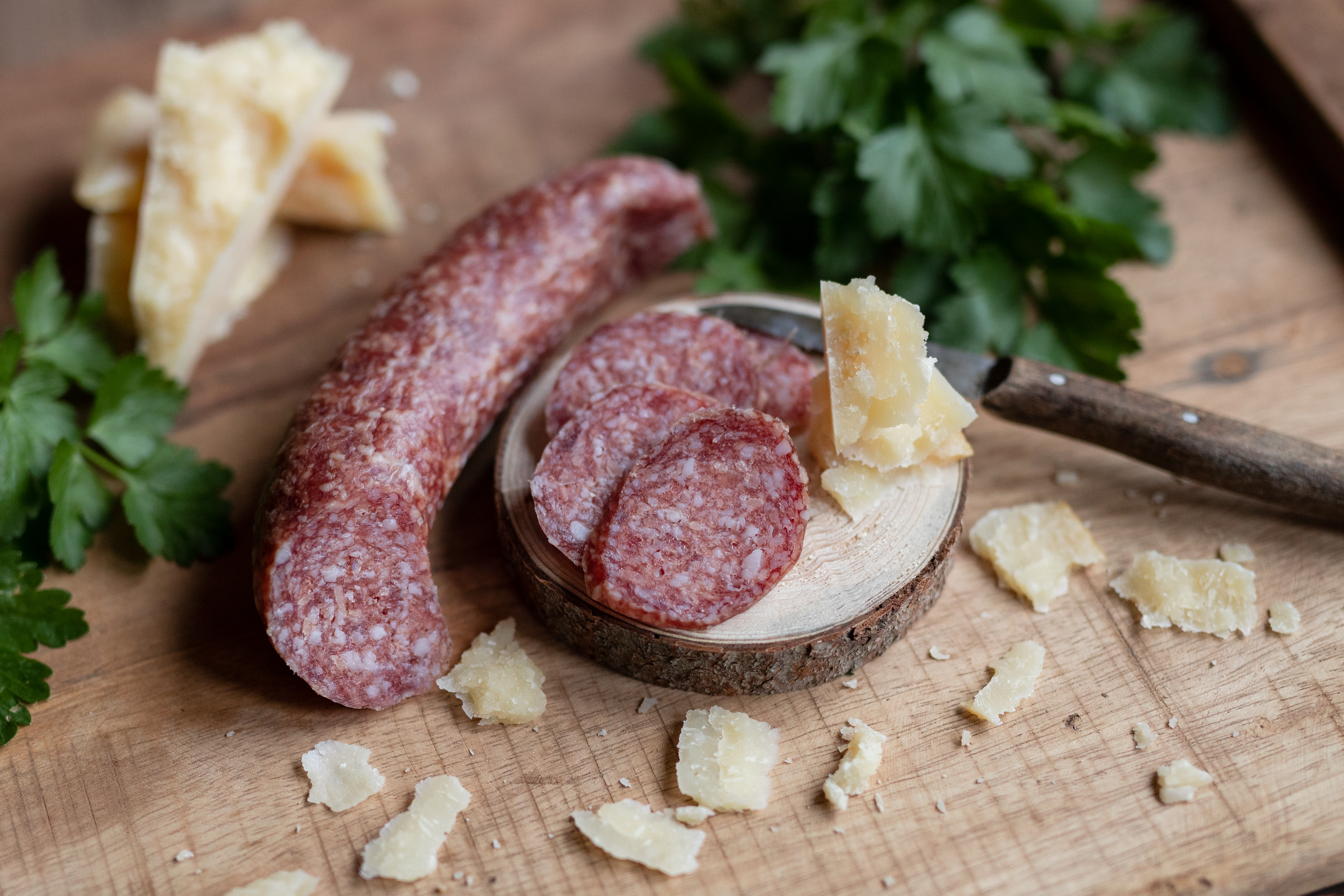 Scheiben Parmesami auf einem runden Brett. Daneben Kräuter, gehobelter Parmesan und die Salami.