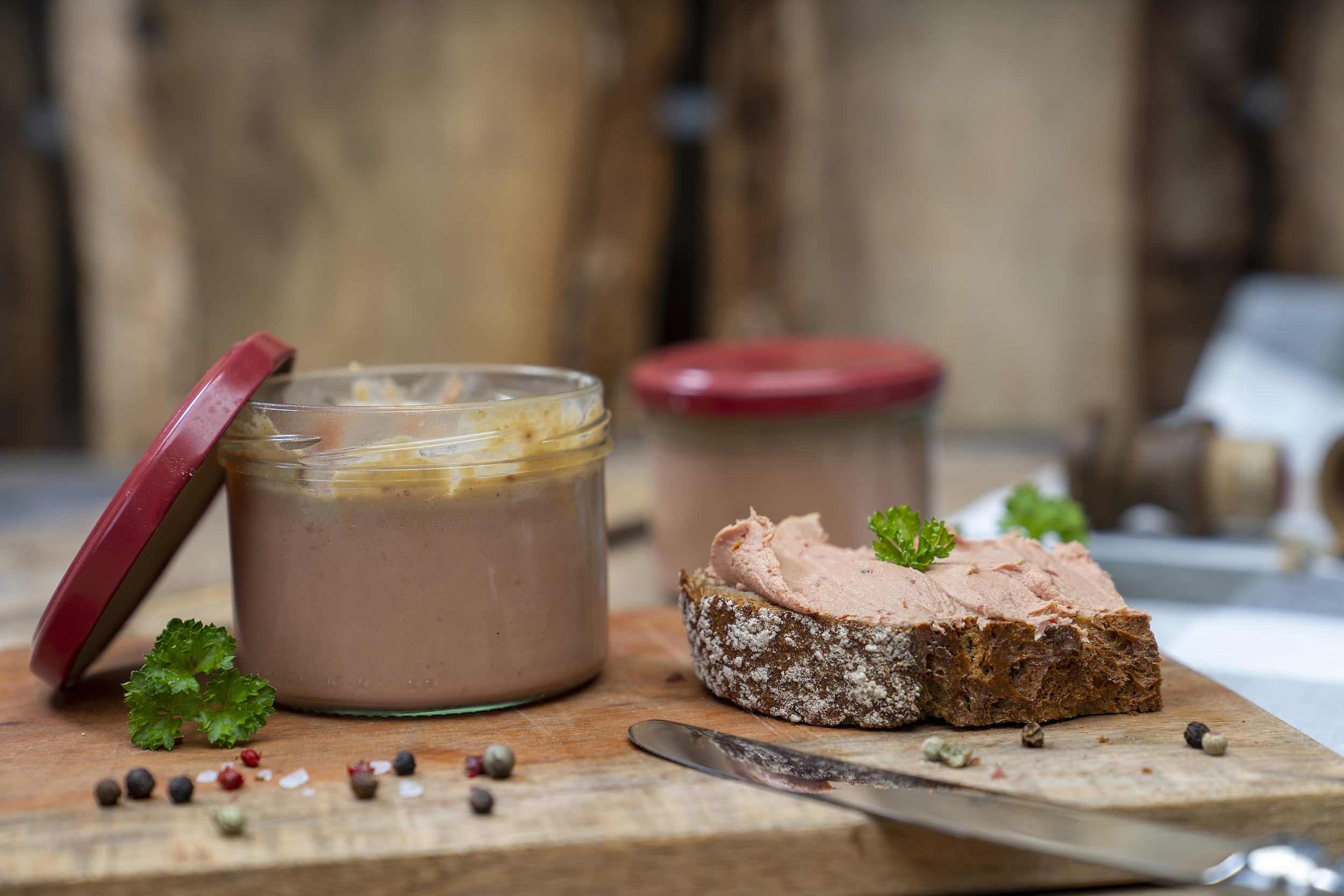 Auf einem Holzbrett ein Glas Tomatenleberwurst. Daneben eine geschmierte Schnitte und Kräuter. Im Hintergrund noch ein Glas.