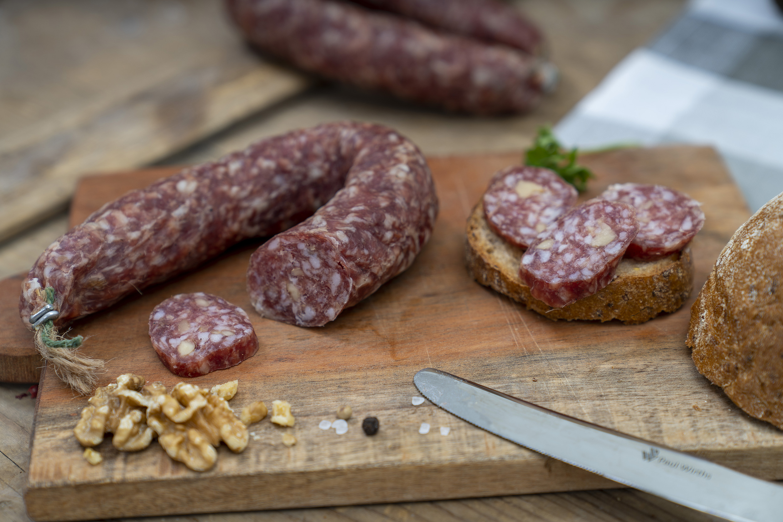 Auf einem Brett eine Walnussschinkenwurst. Daneben Walnüsse, Kräuter und eine Scheibe Brot. Hinten eine weitere Wurst.