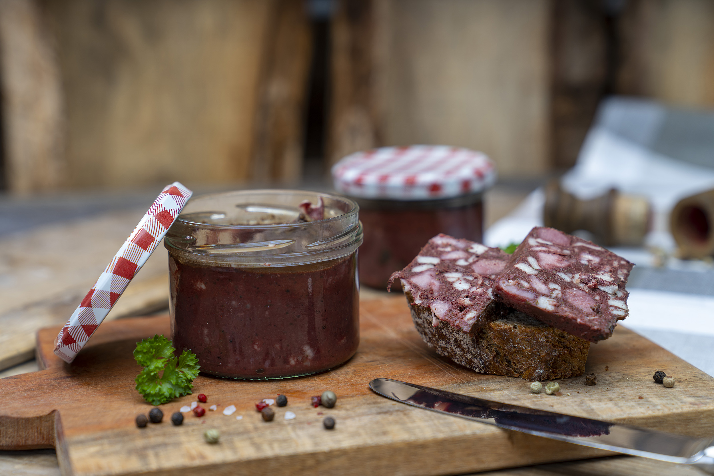 Auf einem Holzbrett steht ein Glas Rotwurst. Daneben eine Scheibe Brot mit Rotwurst Scheiben. Darum Gewürze. Hinten ein weiteres Glas Rotwurst.