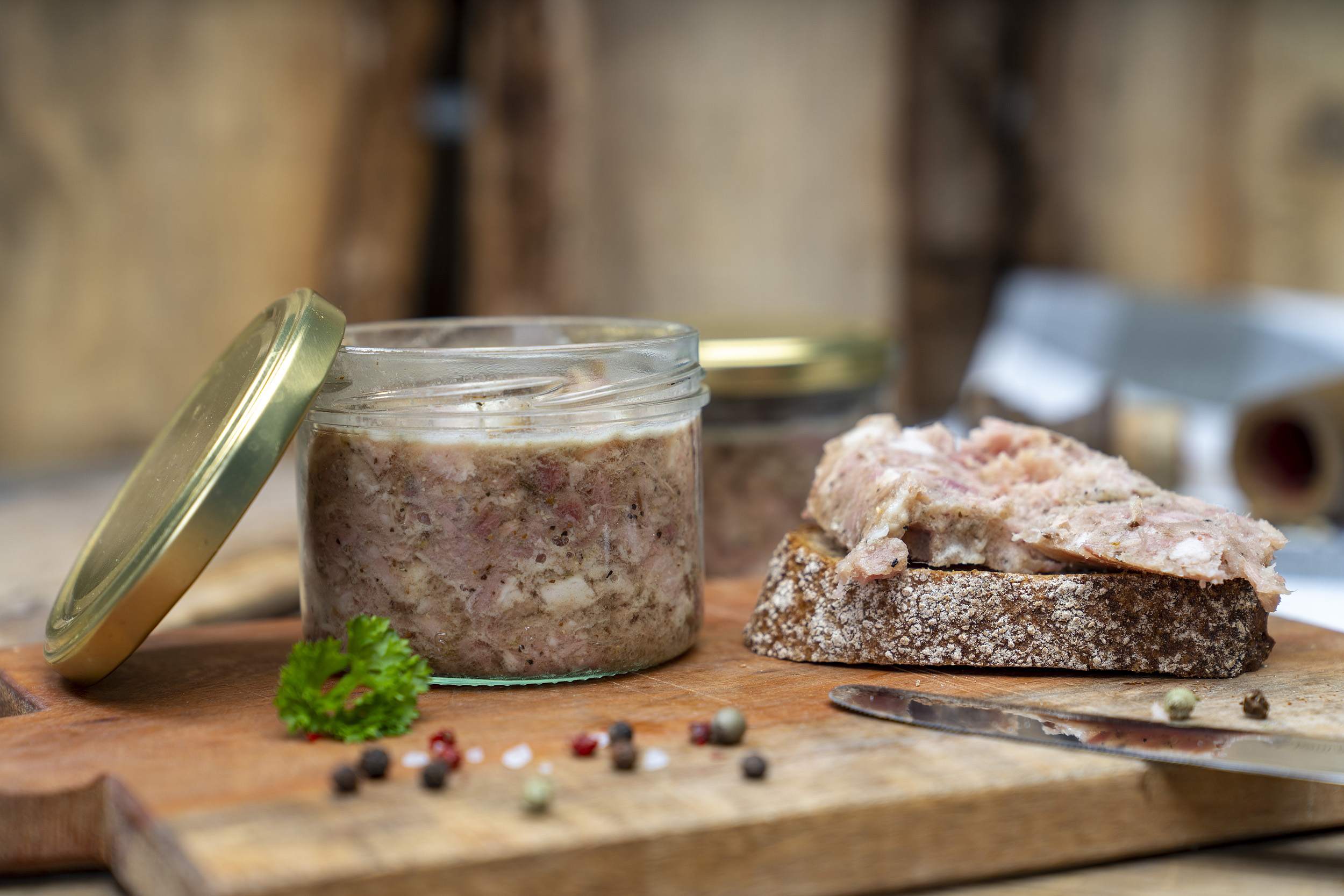 Auf einem Brett ein Glas Sülze. Daneben eine Schnitte mit der Wurst. Drum herum Kräuter und hinten noch ein Glas der Wurst.