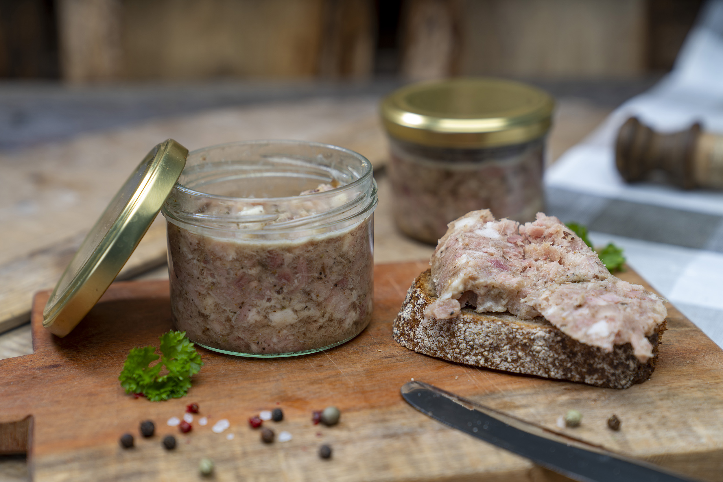 Auf einem Brett ein Glas Sülze. Daneben eine Schnitte mit der Wurst. Drum herum Kräuter und hinten noch ein Glas der Wurst.