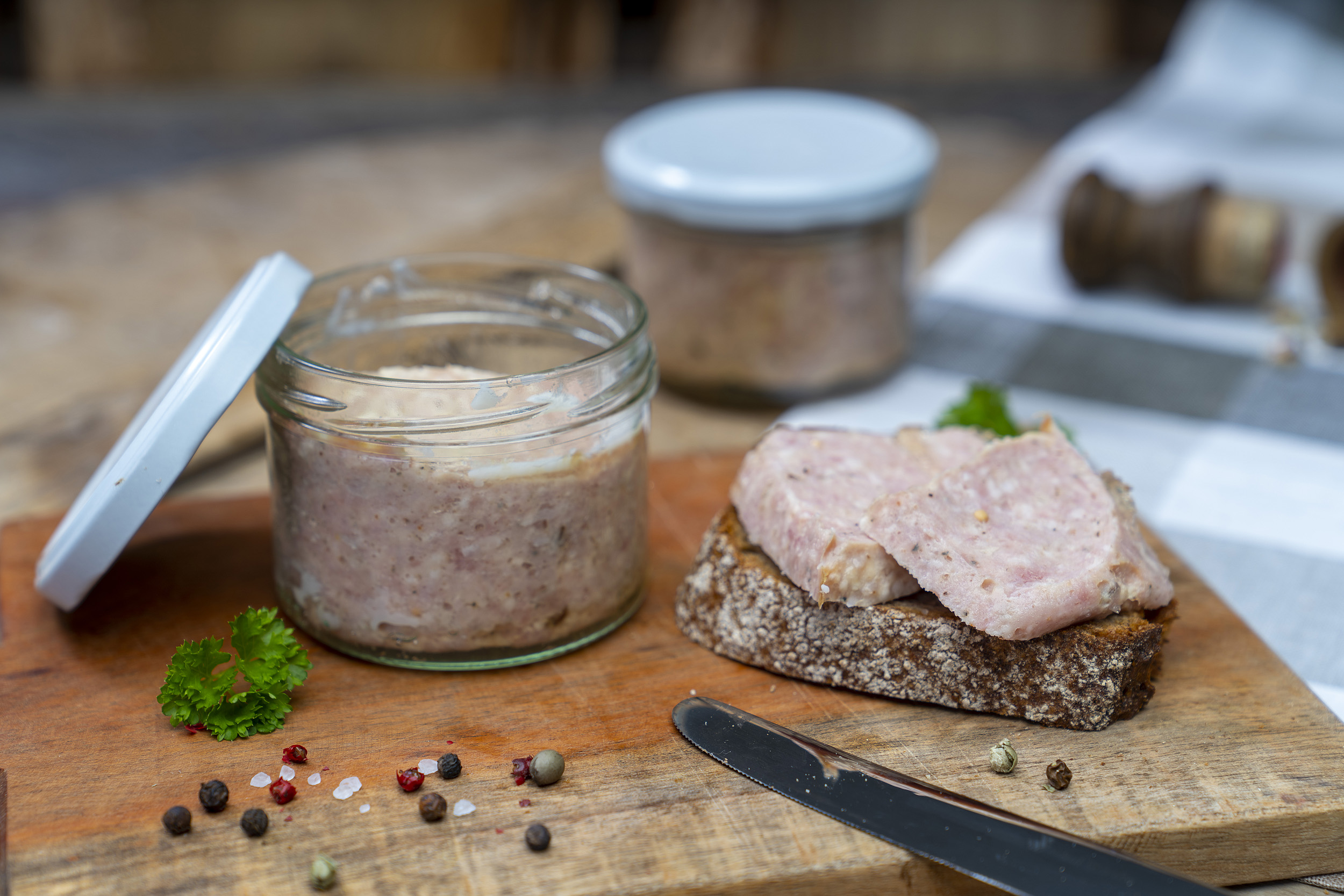 Auf einem Holzbrett ein Glas mit Mettwurst. Daneben eine Schnitte mit Mettwurst, Kräuter und Gewürze. Im Hintergrund steht ein weiteres Glas. Vorn lehnt ein Messer am Brett.