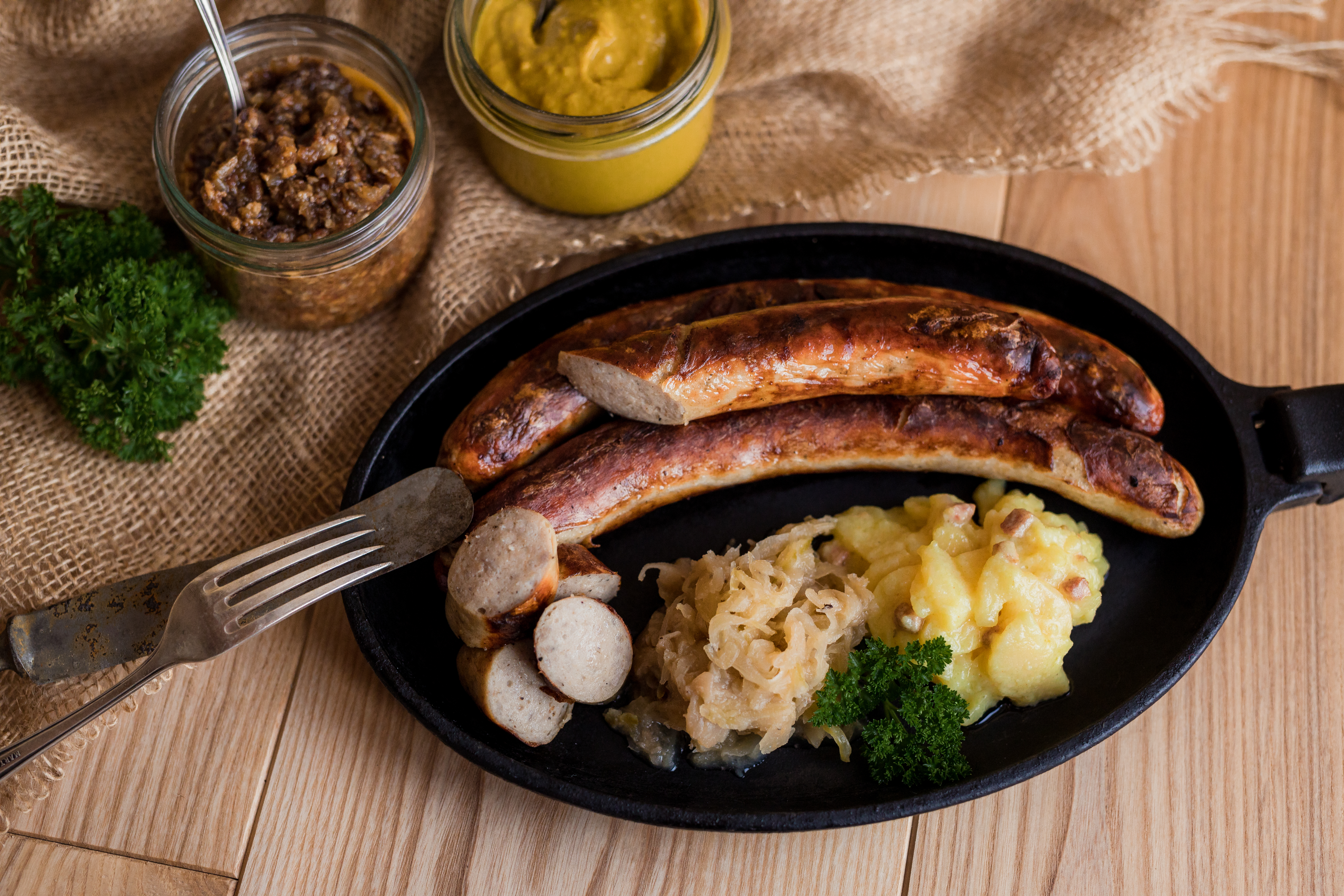 Gebratene Original Thüringer Rostbratwurst in der Pfanne. Mit Sauerkraut und Salatbeilage. Neben der Pfanne Gläser mit Saucen und Kräuter. Untergrund aus Holz.