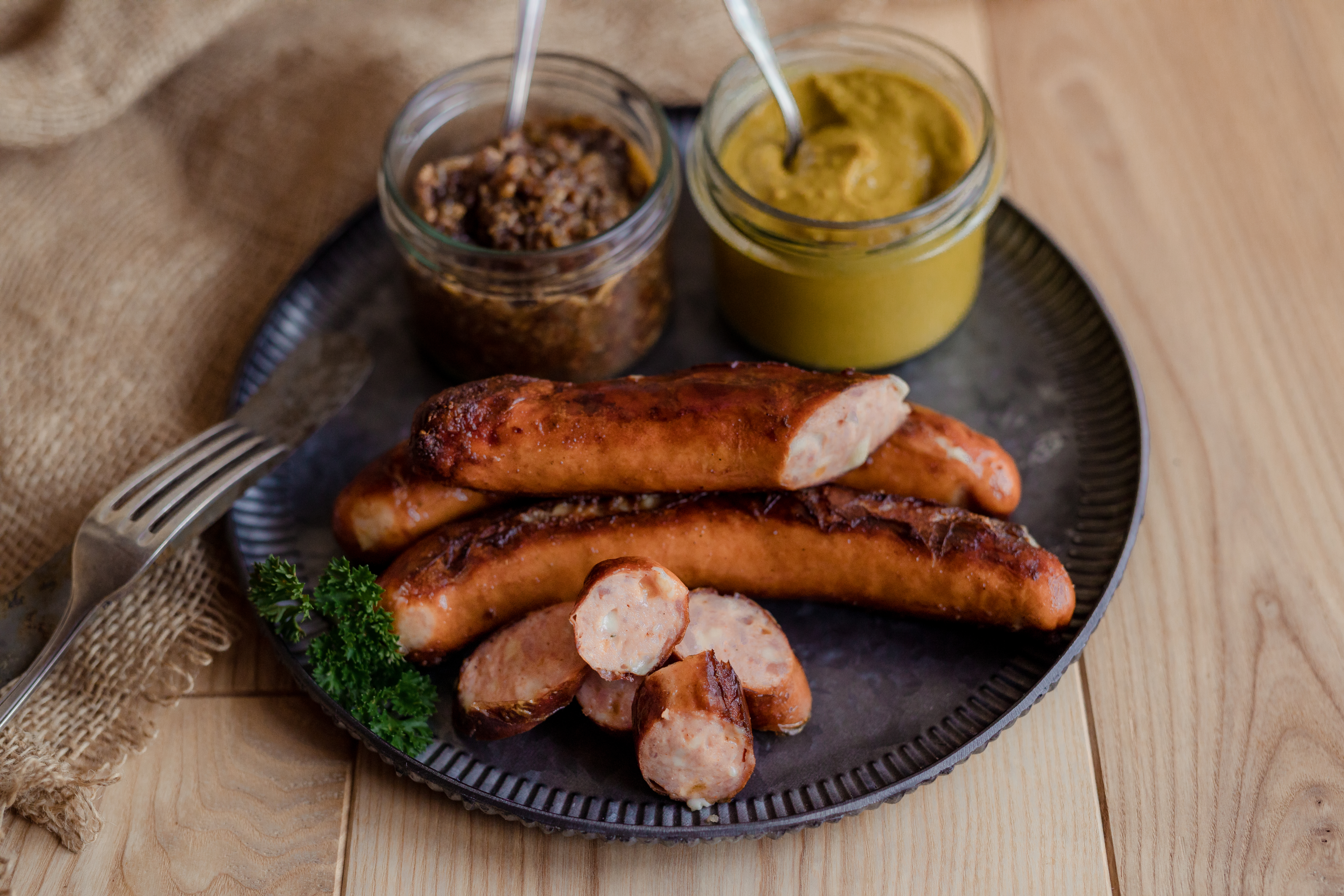Gebratene Käsekrainer auf einem dunklen Teller. Zwei Gläser mit Saucen und Kräuter daneben. Untergrund aus Holz.