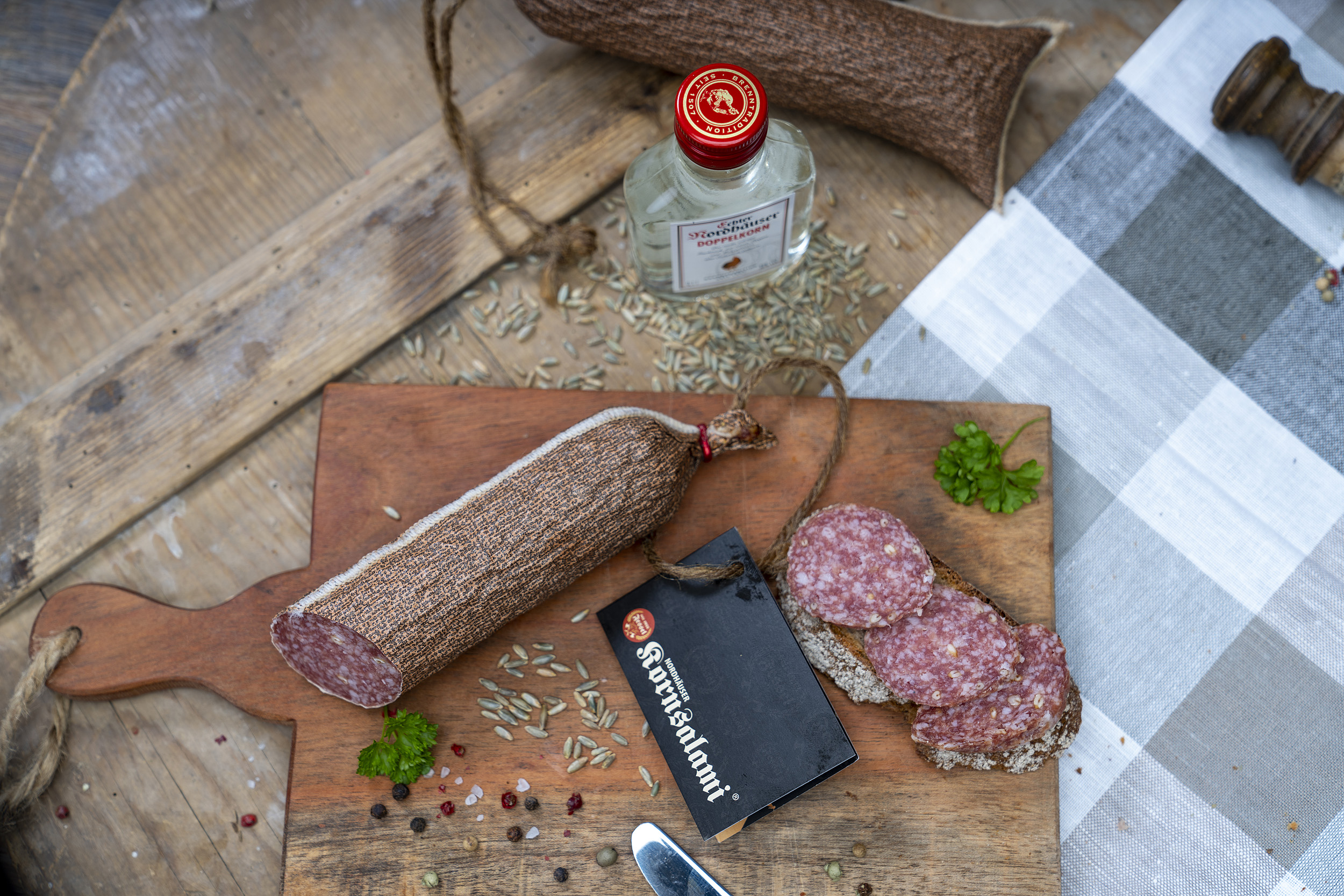 Kornsalami auf einem Holzbrett. Daneben eine Scheibe Brot mit der Salami und Gewürze. Neben dem Brett eine kleine Flasche Echter Nordhäuser Doppelkorn.