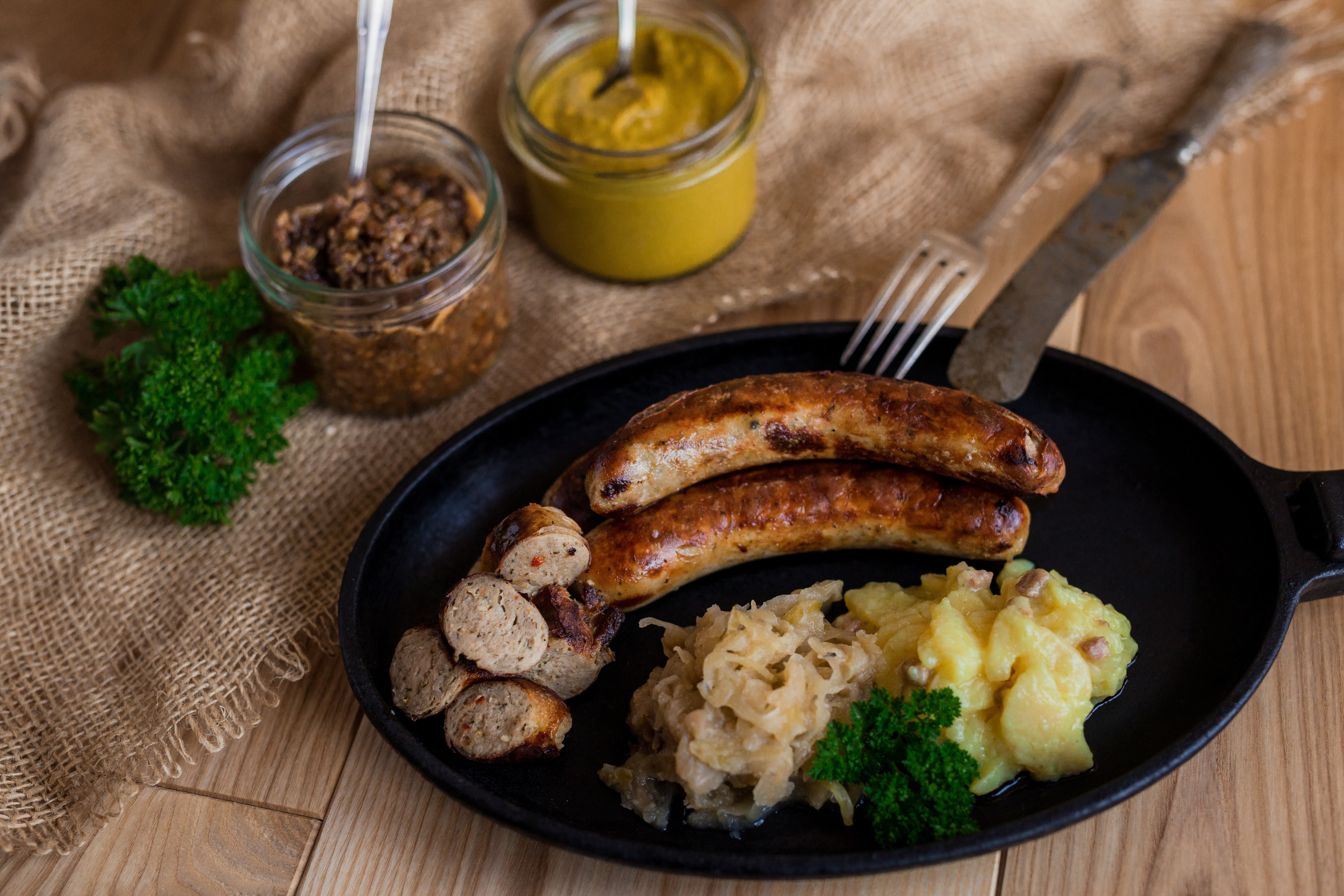 Gebratene Wildbratwurst in der Pfanne. Mit Sauerkraut und Salatbeilage. Neben der Pfanne Gläser mit Saucen und Kräuter. Untergrund aus Holz.