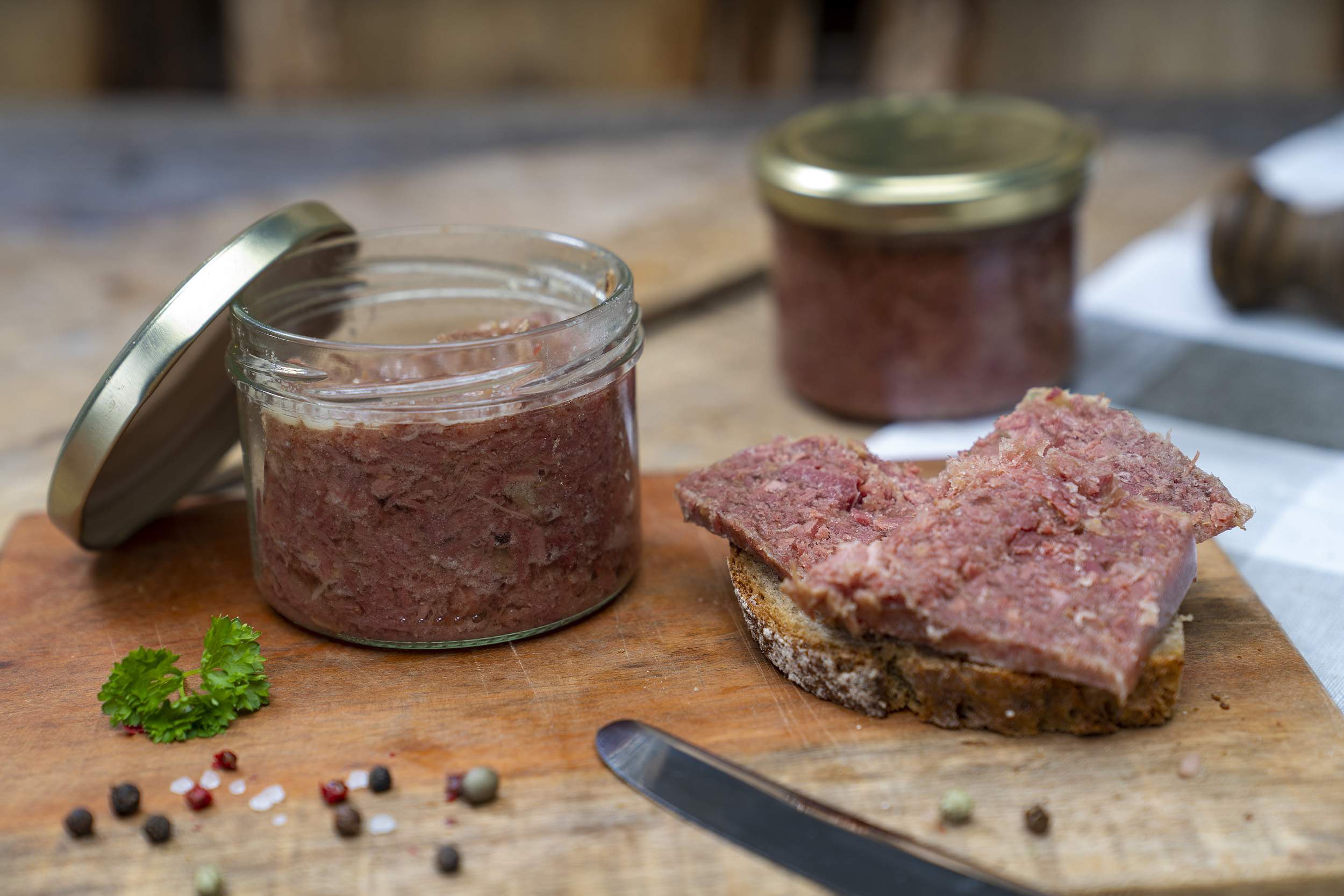 Auf einem Brett ein Glas Corned beef. Daneben ein geschmiertes Brot mit Corned beef. Daneben Gewürze, Kräuter und ein Messer. Im Hintergrund ein weiteres Glas.