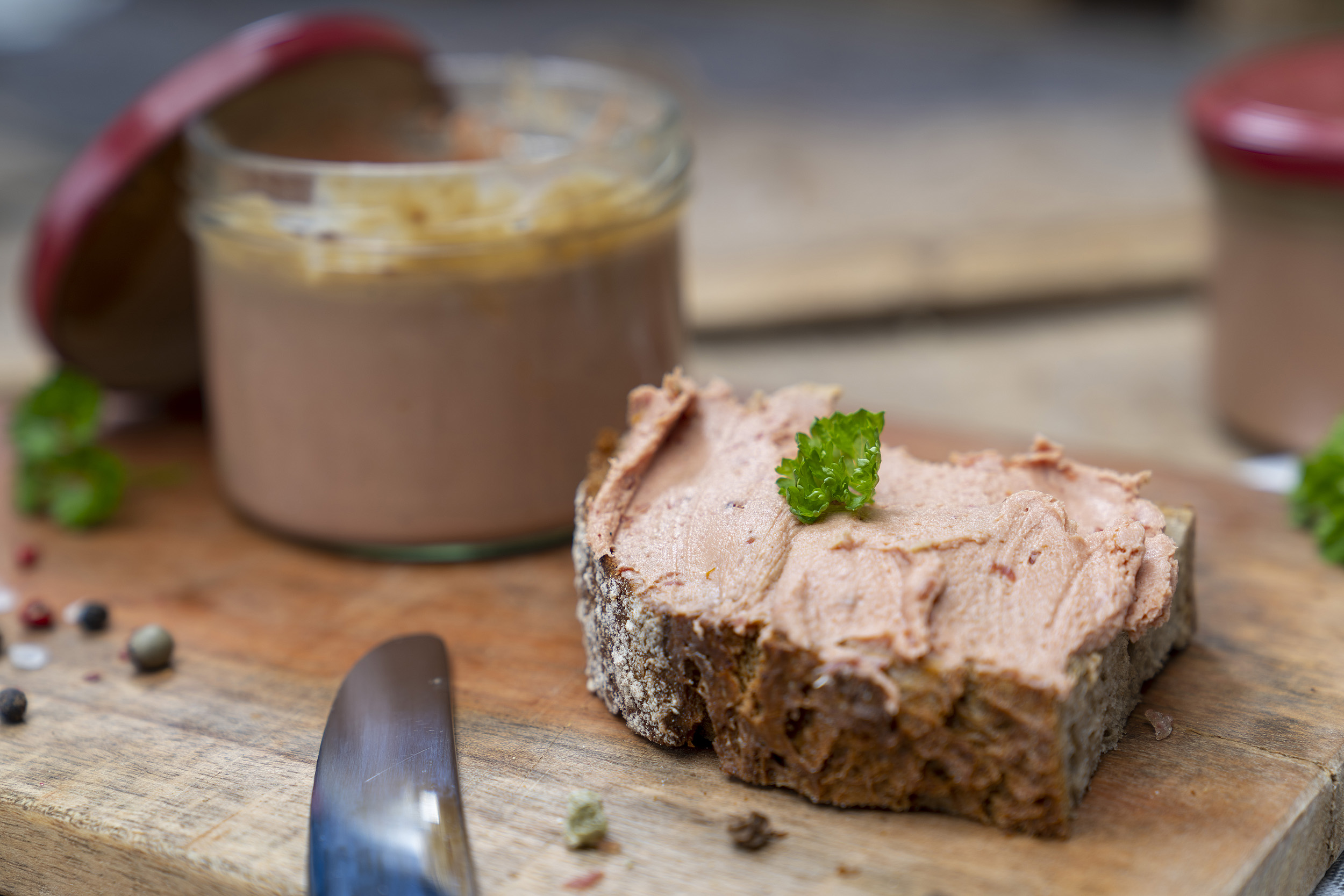 Auf einem Holzbrett ein Glas Tomatenleberwurst. Daneben eine geschmierte Schnitte und Kräuter. Im Hintergrund noch ein Glas.