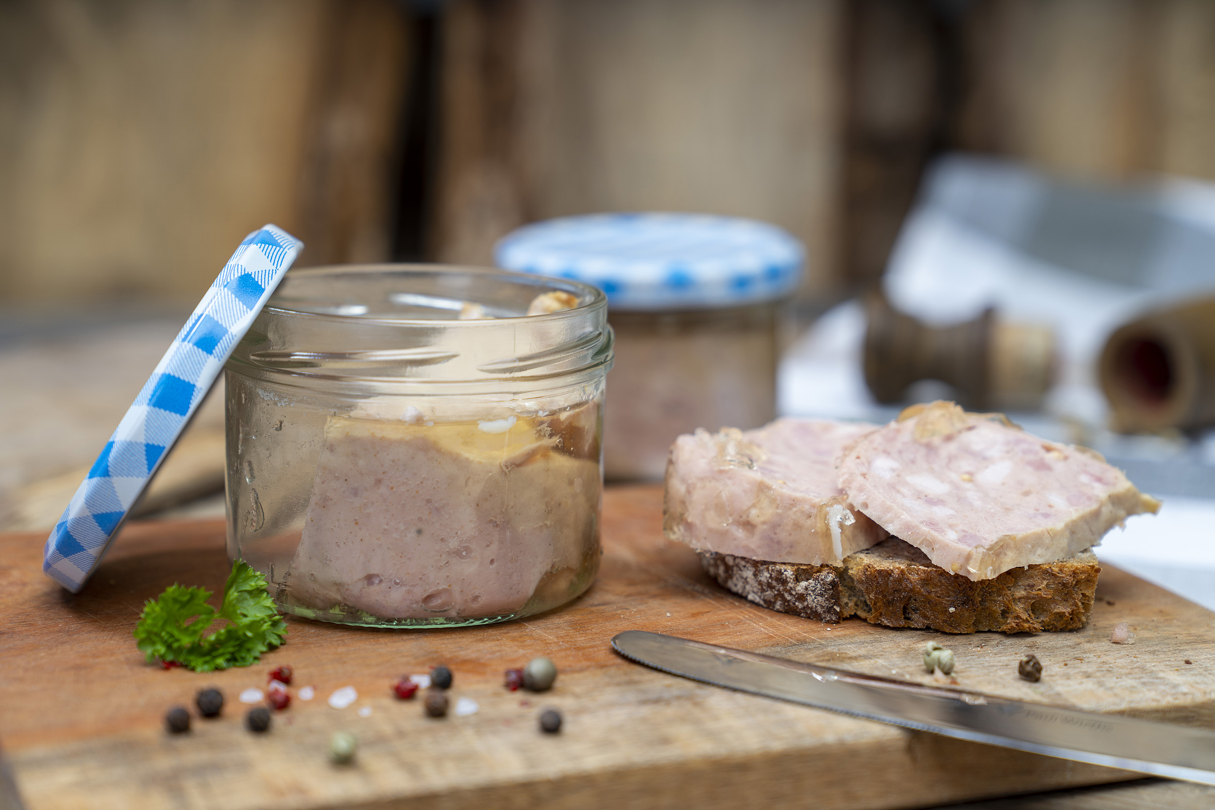 Auf einem Holzbrett ein Glas Jagdwurst. Daneben eine Schnitte mit der Jagdwurst. Neben dem Glas Gewürze, Kräuter und ein Messer. Im Hintergrund ein weiteres Glas Jagdwurst.