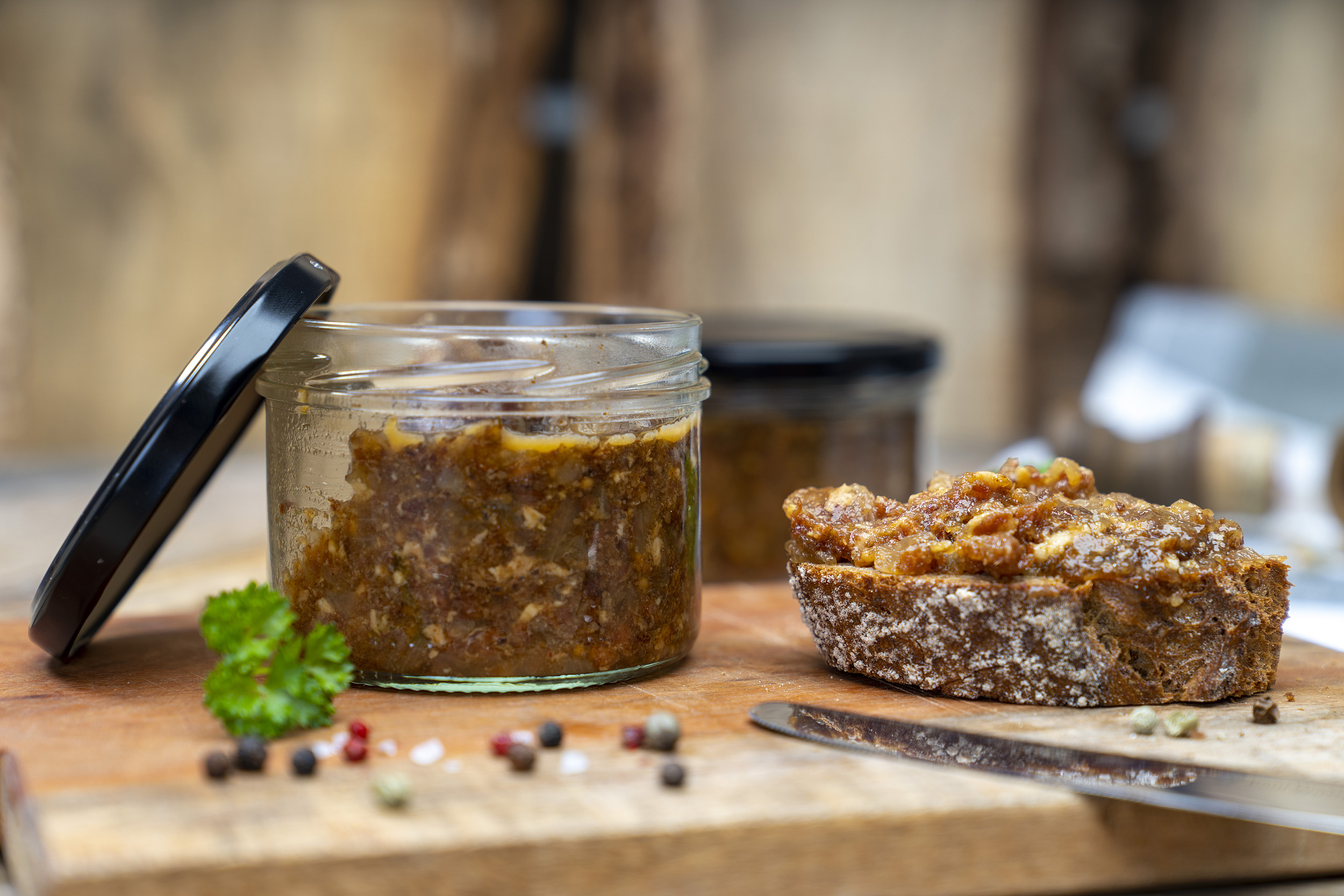 Auf einem Holzbrett steht ein Glas mit Bacon Jam. Daneben Gewürze, Kräuter und ein geschmiertes Brot mit der Marmelade. Im Hintergrund ein weiteres Glas. Vorn ein Messer am Brett.