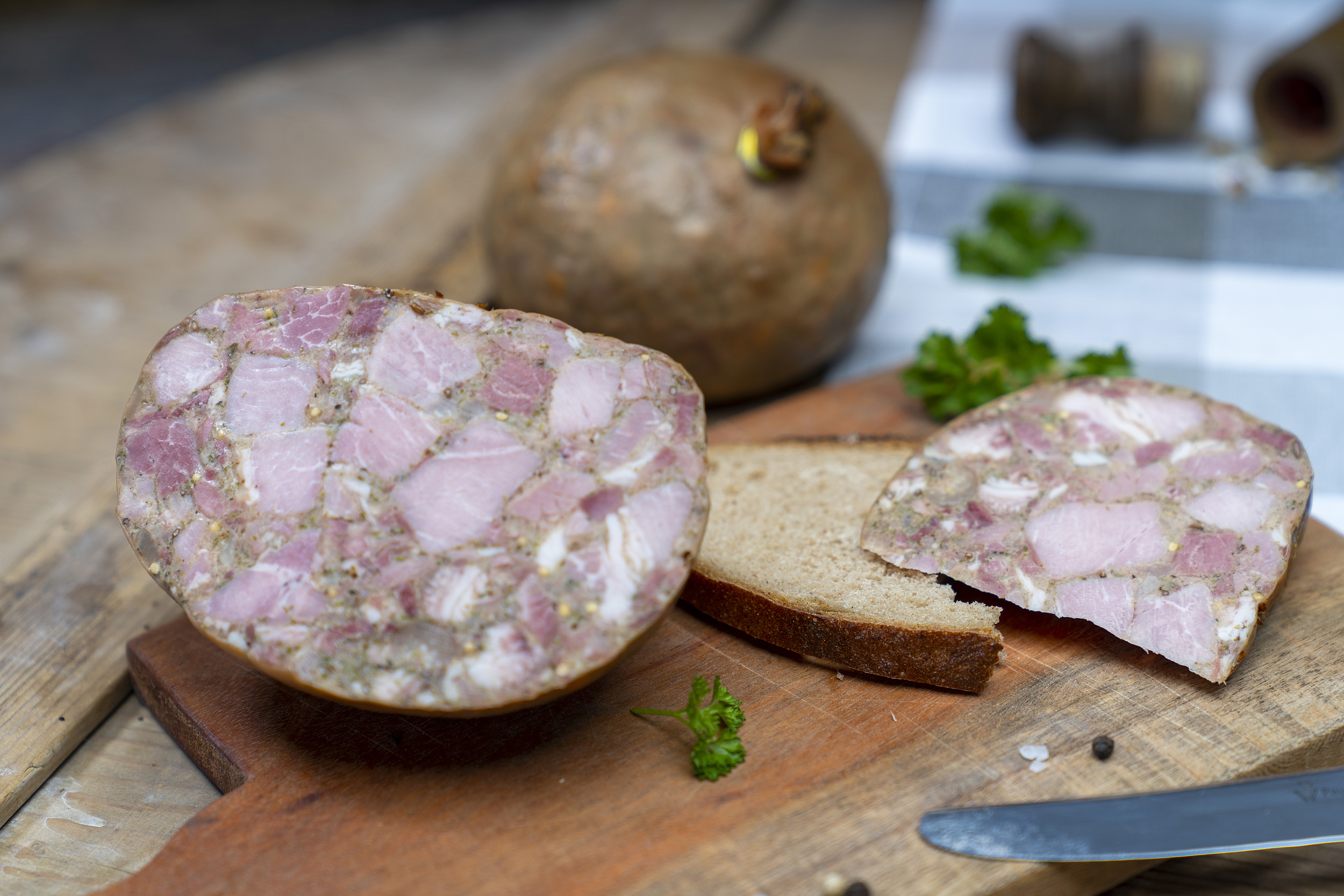 Eine Scheibe mit Sülze auf einem Brett. Daneben die angeschnittene Blase der Wurst und Gewürze. Hinten eine weitere Blase.